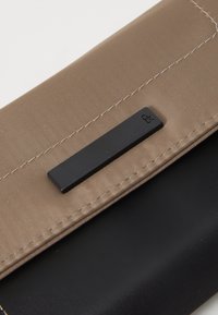 Portofel din material textil maro cu un accent de hardware rectangular negru. Prezintă cusături texturate și o suprafață interioară netedă.