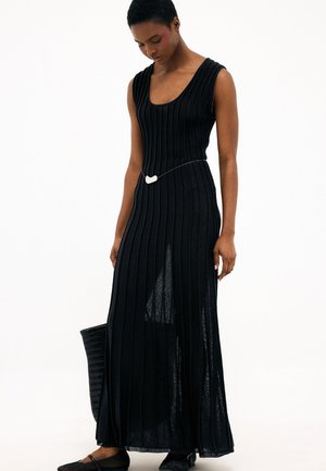 Žena má na sebe čierne rebrované maxi šaty bez rukávov s bočným rozparkom, tenkým opaskom, čierne baleríny a drží čiernu tkanú tašku.