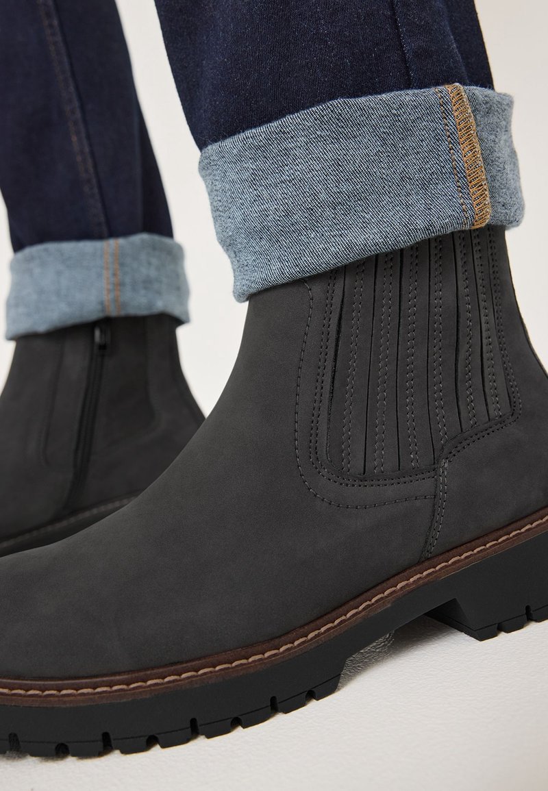 Bottines Chelsea gris foncé avec panneaux latéraux élastiques et coutures marron, portées avec un jean en denim bleu retroussé.