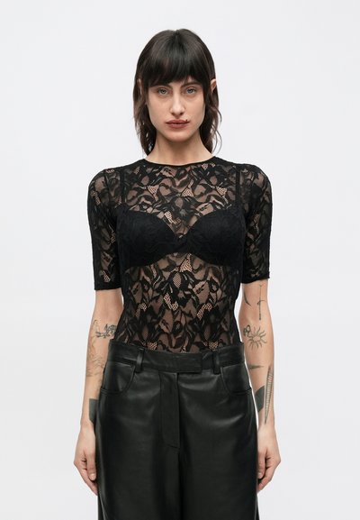Body in pizzo nero con maniche corte e motivo floreale, indossato sopra un reggiseno nero, abbinato a pantaloni in pelle a vita alta.