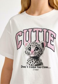 Hvid bomulds T-shirt med et grafisk motiv af en plettet killing, med lyserøde accenter og teksten "CUTIE" ovenfor og "Kom ikke for tæt på... Jeg bider!" nedenfor.
