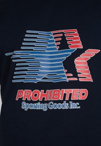 Logotipo estilizado de tiburón azul y rojo sobre texto en negrita rojo "PROHIBITED" y azul "Sporting Goods Inc." sobre fondo de tela oscura.