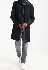 Man in een zwarte trenchcoat over een blauw geruit overhemd, grijze jeans en witte sneakers, die naar voren loopt tegen een witte achtergrond.