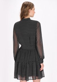 Zwart mini-jurk met doorzichtige mouwen, een getailleerde taille en een gerimpelde zoom; heeft een geometrisch patroon in het gehele ontwerp. Achteraanzicht weergegeven.