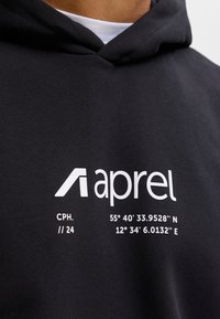 Kapucar s logotipom "aprel" in besedilom, ki prikazuje koordinate 55°40'33.9528''N 12°34'6.0132''E ter "CPH. // 24" na črni tkanini.