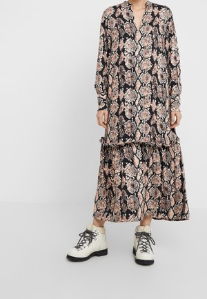 Kobieta ubrana w długą sukienkę w wężowy wzór z falbanami, do której dobrała białe, wiązane botki na kostkę, na jednolitym tle.