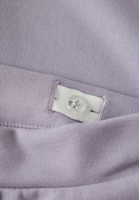 Violetti kangas, jossa sileä tekstuuri, valkoinen etiketti ja selkeä nappi ommeltuna. Saumat ovat siistit ja tasaiset.