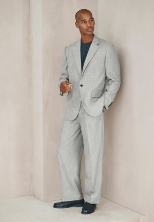 Man in light gray suit and dark shirt stands against beige wall, one hand in pocket and the other slightly raised, wearing black shoes.