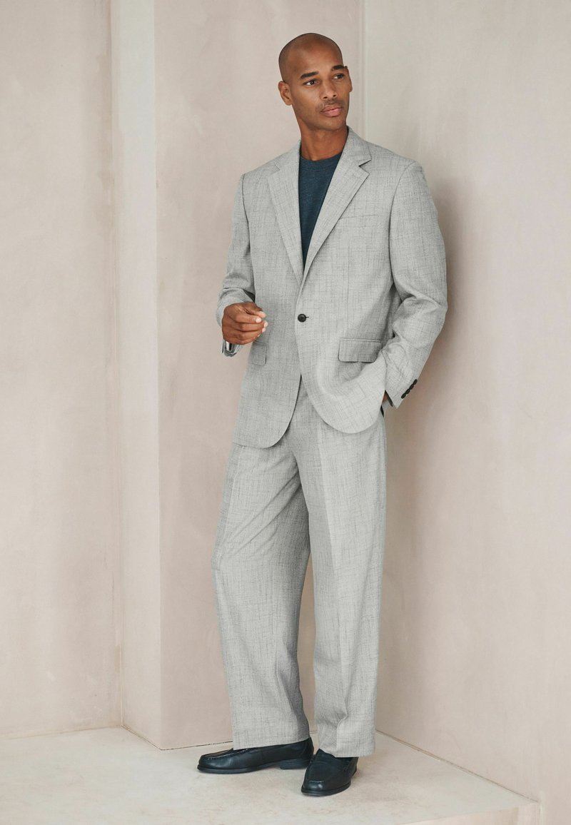 Homme en costume gris clair et chemise foncée, debout contre un mur beige, une main dans la poche et l'autre légèrement levée, portant des chaussures noires.