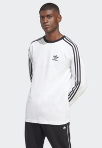 Biela dlhá mikina s čiernymi akcentmi, so tromi čiernymi pruhmi na rukávoch a logom Adidas na hrudi.