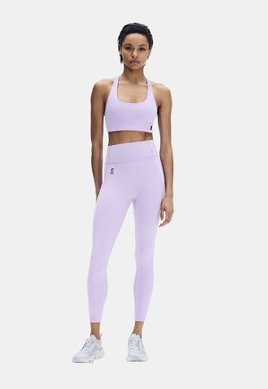 Femme debout portant un soutien-gorge de sport violet clair et un legging assorti avec des baskets blanches sur fond uni.