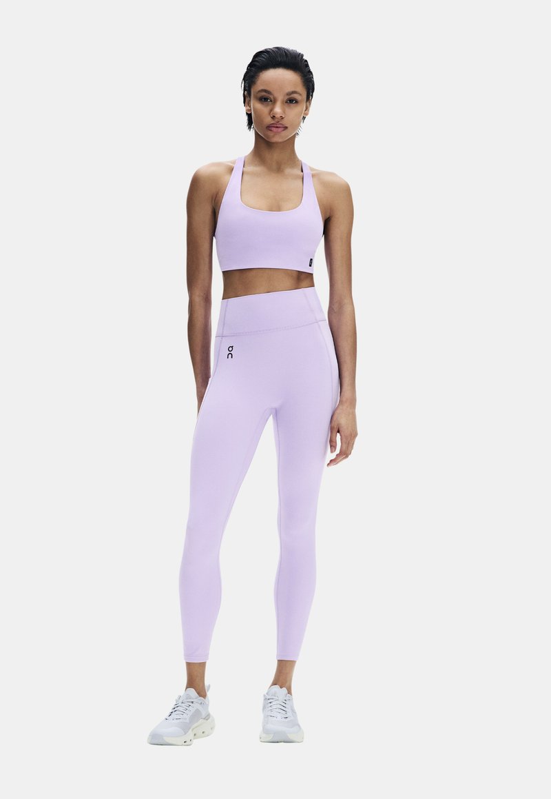 Femme debout portant un soutien-gorge de sport violet clair et un legging assorti avec des baskets blanches sur fond uni.