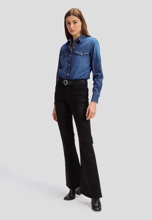 Femme debout portant une chemise en jean bleu à boutons, un pantalon noir évasé avec ceinture noire et des bottes à talons noires sur un fond gris clair.