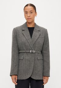 Blazer cinza com padrão pied-de-poule, cinto ajustado, lapelas largas e dois bolsos frontais. Tecido texturizado com ombros estruturados.