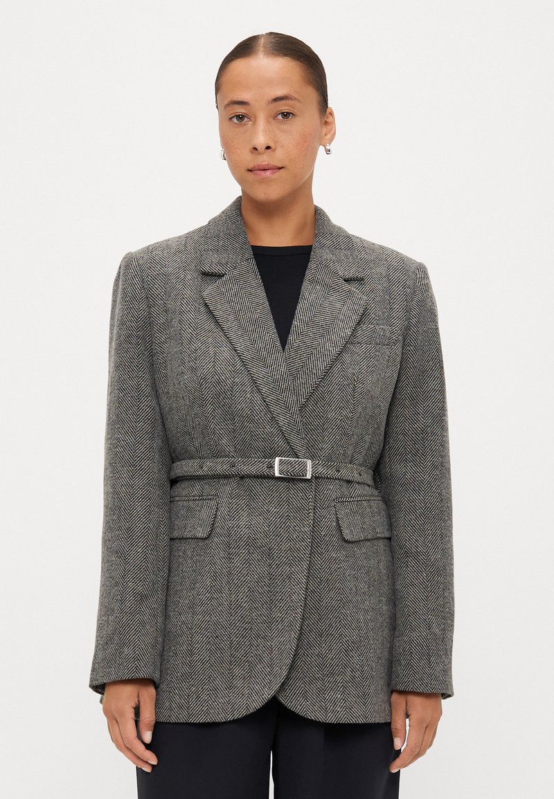 Blazer cinza com padrão pied-de-poule, cinto ajustado, lapelas largas e dois bolsos frontais. Tecido texturizado com ombros estruturados.