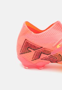 Helles pinkes Fußballschuh mit einem gestrickten Obermaterial, strukturierten Akzenten und transparenten Stollen. Mit geometrischen Mustern in Schwarz und Orange.