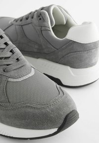 Grå sneakers gjorda av mesh och mocka; har rundad tå, snörningsdesign, vit detalj vid hälen och texturerade ytor.