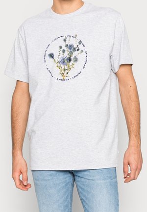 Persona che indossa una maglietta grigio chiaro con una grafica circolare di fiori di cardo blu e un testo che recita "Amore per il mondo e non fare del male, crea."