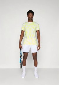 Sport T-shirt met korte mouwen in geel en grijs patroon, gecombineerd met witte korte broek en sportschoenen, met een tennisracket in de hand. Eenvoudig ontwerp.