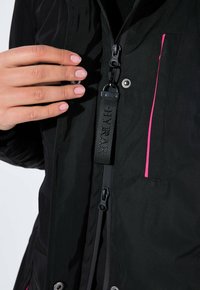 Musta takki, jossa on sileä pinta, vaaleanpunaisella vetoketjulla ja näkyvällä tarralla, jossa lukee "HYBRAR." Käsi sijoitettu lähelle vetoketjun vetämistä.