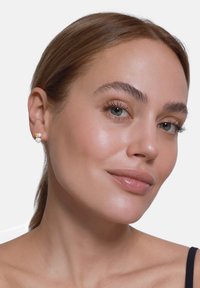 Boucles d'oreilles en or et perles, avec un design texturé, présentant une perle blanche centrale et de petites touches dorées. La peau est lisse et claire.
