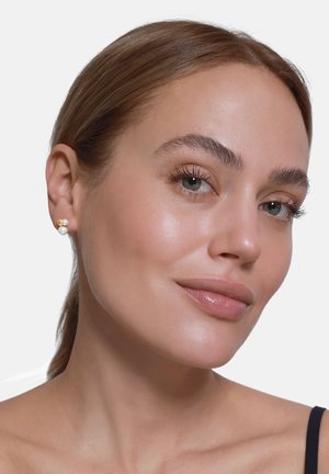 Boucles d'oreilles en or et perles, avec un design texturé, présentant une perle blanche centrale et de petites touches dorées. La peau est lisse et claire.
