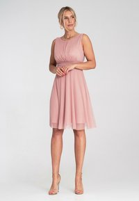 Różowa sukienka bez rękawów z dopasowanym gorsetem, fałdami w talii i zwiewną, przezroczystą spódnicą. Modelka nosi minimalistyczne obcasy i biżuterię.