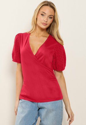 Blondynka w czerwonym topie z dekoltem w serek, z teksturowanymi bufkami i jasnoniebieskich dżinsach, stojąca na jasnym, jednolitym tle.