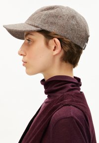 Grijze, gestructureerde wollen baseballpet met een gebogen klep en een embleem aan de zijkant, gedragen door een persoon in een bordeauxrode coltrui. Profielweergave.