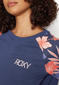 Femme aux cheveux bouclés portant un sweat-shirt bleu avec des manches à motifs floraux et le mot « ROXY » imprimé sur la poitrine.