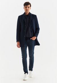 Granatowy płaszcz wełniany z blendą o dopasowanym kroju, z przodem na jeden guzik, wyciętymi klapami i guzikami, noszony na wzorzystym swetrze i dżinsach.