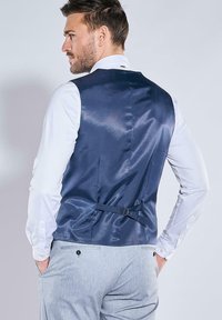 Marineblaues Satinweste mit glatter Textur, verstellbarer Schnalle auf der Rückseite, getragen über einem weißen Hemd, kombiniert mit grauen Hosen.