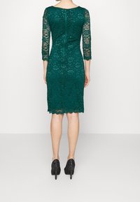 Femme portant une robe en dentelle vert foncé longueur genou avec des manches trois-quarts et des talons hauts noirs pailletés, de dos à la caméra.