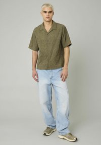 Chemise olive à manches courtes avec motif en relief de feuilles, associée à un jean bleu clair à coupe ample et des baskets grises avec des accents noirs.