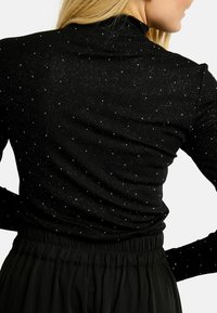 Kobieta ubrana w czarną bluzkę z długim rękawem z białym kropkowanym wzorem oraz czarne spodnie z marszczonym paskiem, widziana od tyłu i z boku.