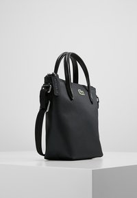 Bolso sintético negro con superficie texturizada, asas dobles, correa desmontable y un pequeño logo verde. Cierre de cremallera y detalles en hardware plateado.