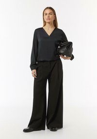 Schwarze Seidenbluse mit V-Ausschnitt und puffärmeligen Ärmeln, kombiniert mit weit geschnittenen schwarzen Hosen und einer strukturierten schwarzen Handtasche.