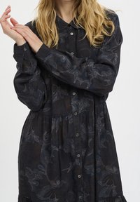 Kvinna med långt blont hår som bär en mörk knäppt klänning med subtila blommönster, justerar ärmen på vänster arm mot en neutral vit bakgrund.