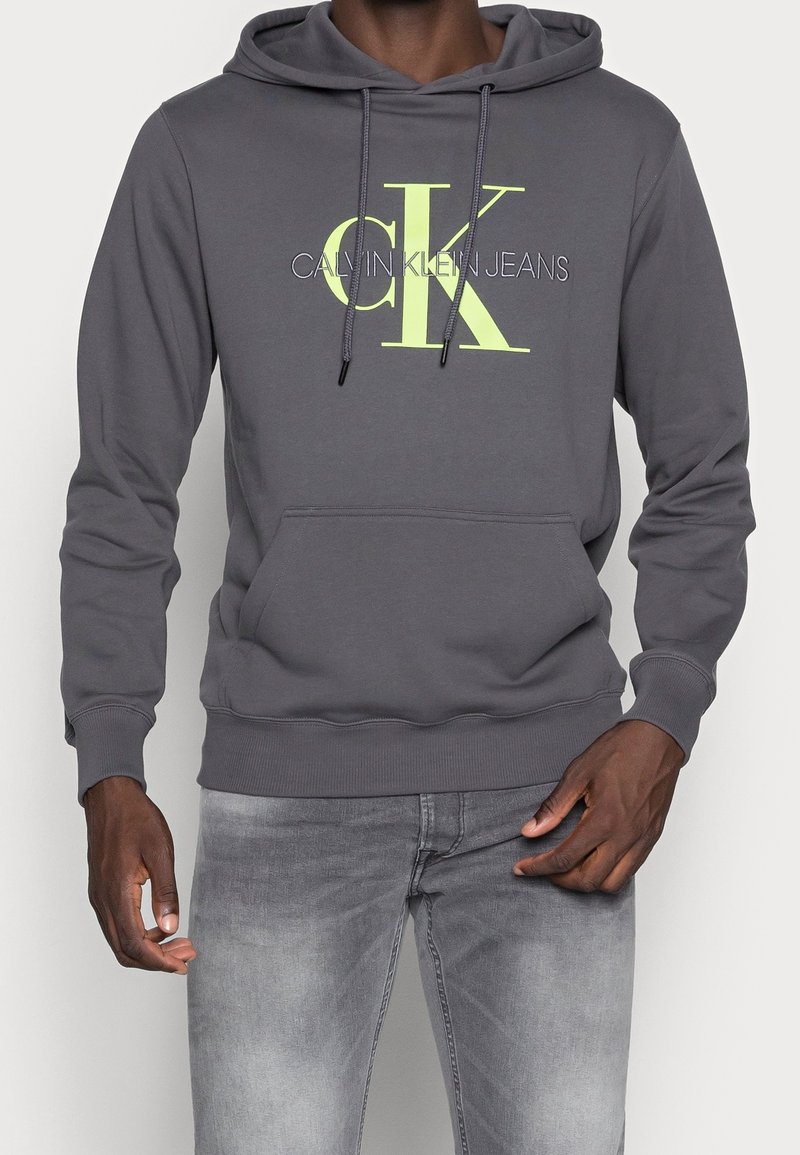 Homme portant un sweat à capuche gris foncé avec un grand texte jaune "CK" et "Calvin Klein Jeans" et un jean gris délavé, debout les mains près des poches.