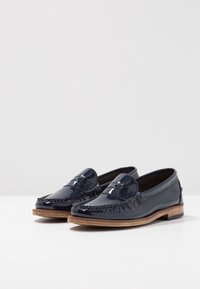 Mocassins en cuir verni bleu marine avec un design de moccasin, des détails à franges et une semelle en bois naturel. Texture lisse avec un éclat subtil.
