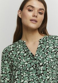 Svart blus med grönt blommönster, V-ringning och knapprad fram. Tyget har en mjuk textur och ett lätt utseende.