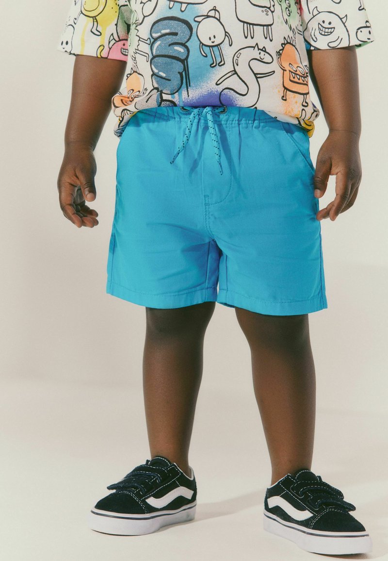 Enfant portant un short bleu vif, des baskets noires et blanches, et un t-shirt coloré avec des personnages de dessins animés, debout devant un fond uni.