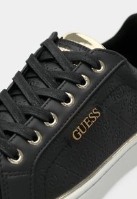 Baskets en cuir noir avec motif embossé, lacets noirs passant par des œillets dorés et logo "GUESS" doré sur le côté au-dessus de la semelle blanche.