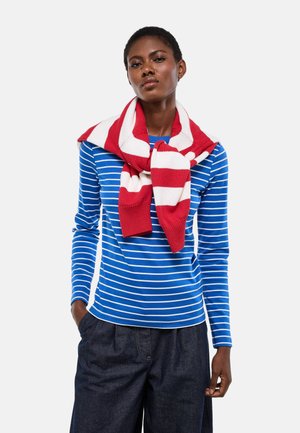 Femme portant une chemise à manches longues rayée bleu et blanc, un pantalon ample foncé, et un pull rayé rouge et blanc posé sur ses épaules.