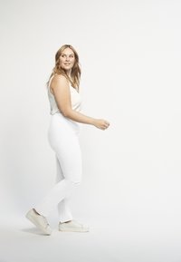 Witte jeans gecombineerd met een licht, geribbeld hemdje. Het model draagt witte sneakers en heeft golvend, schouderlang haar. Eenvoudig, strak ontwerp.
