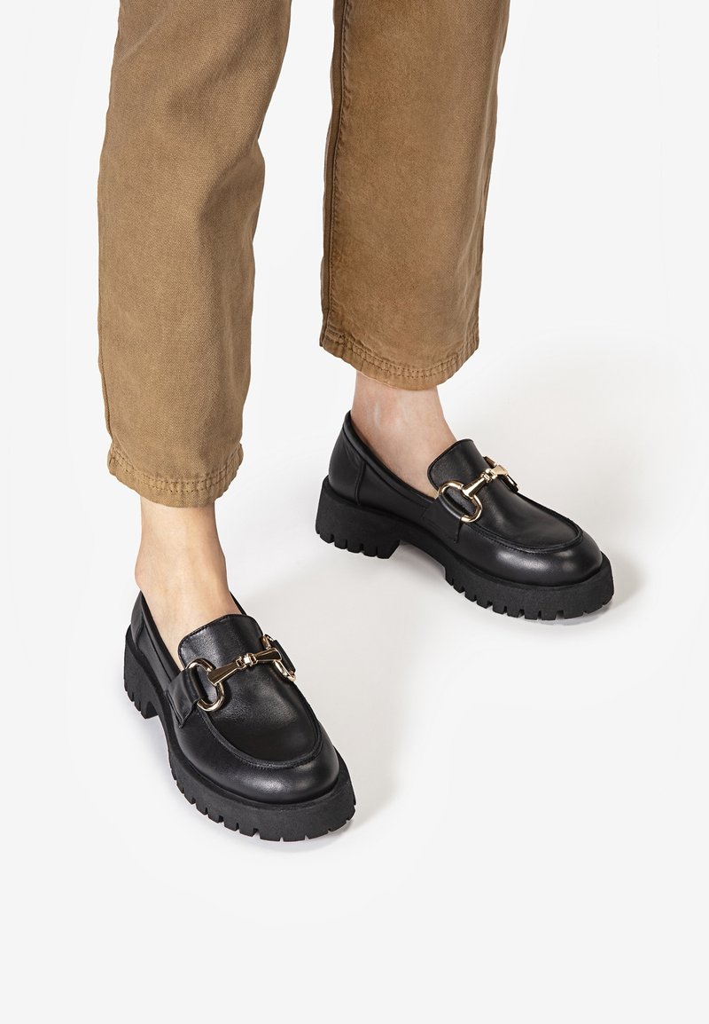 Czarne skórzane mokasyny z grubymi podeszwami i złotymi metalowymi klamrami noszone z przyciętymi brązowymi spodniami na bosych stopach.