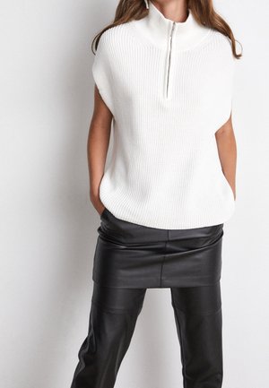 Femme portant un pull sans manches en tricot blanc avec une fermeture éclair à l'avant et une mini-jupe en cuir noire par-dessus un pantalon en cuir noir, les mains dans les poches.