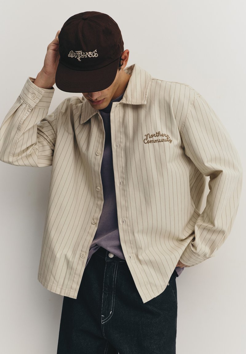 Chemise beige à rayures avec boutons et broderie "Northern Community". Casquette foncée avec logo "Outlaws" en écriture cursive. Jean noir et chemise violette en dessous.