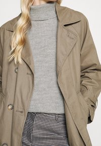 Beige trenchcoat i lättviktsmaterial med notched krage och knäppning, lager på lager över en grå ribbad polotröja.