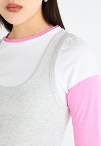Top de tirantes gris con textura de canalé, superpuesto a una camisa de manga larga blanca con cuello rosa. Textura suave y diseño sencillo, centrado en colores contrastantes.